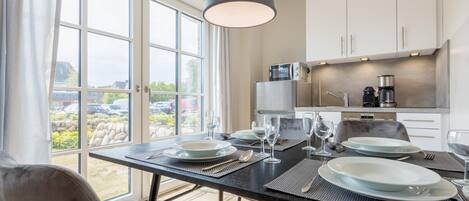 Un comedor moderno con una mesa preparada para cuatro, una cocina con electrodomésticos de acero inoxidable y vistas a un jardín a través de amplios ventanales.