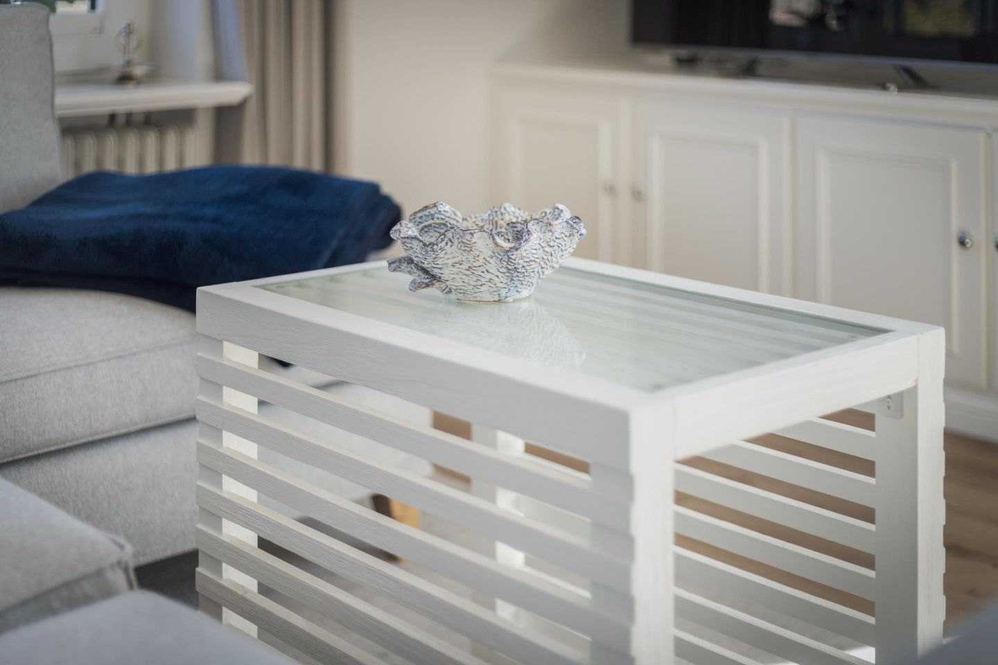 Une petite table d’appoint blanche, avec un plateau en verre et un bol décoratif en céramique posé dessus.