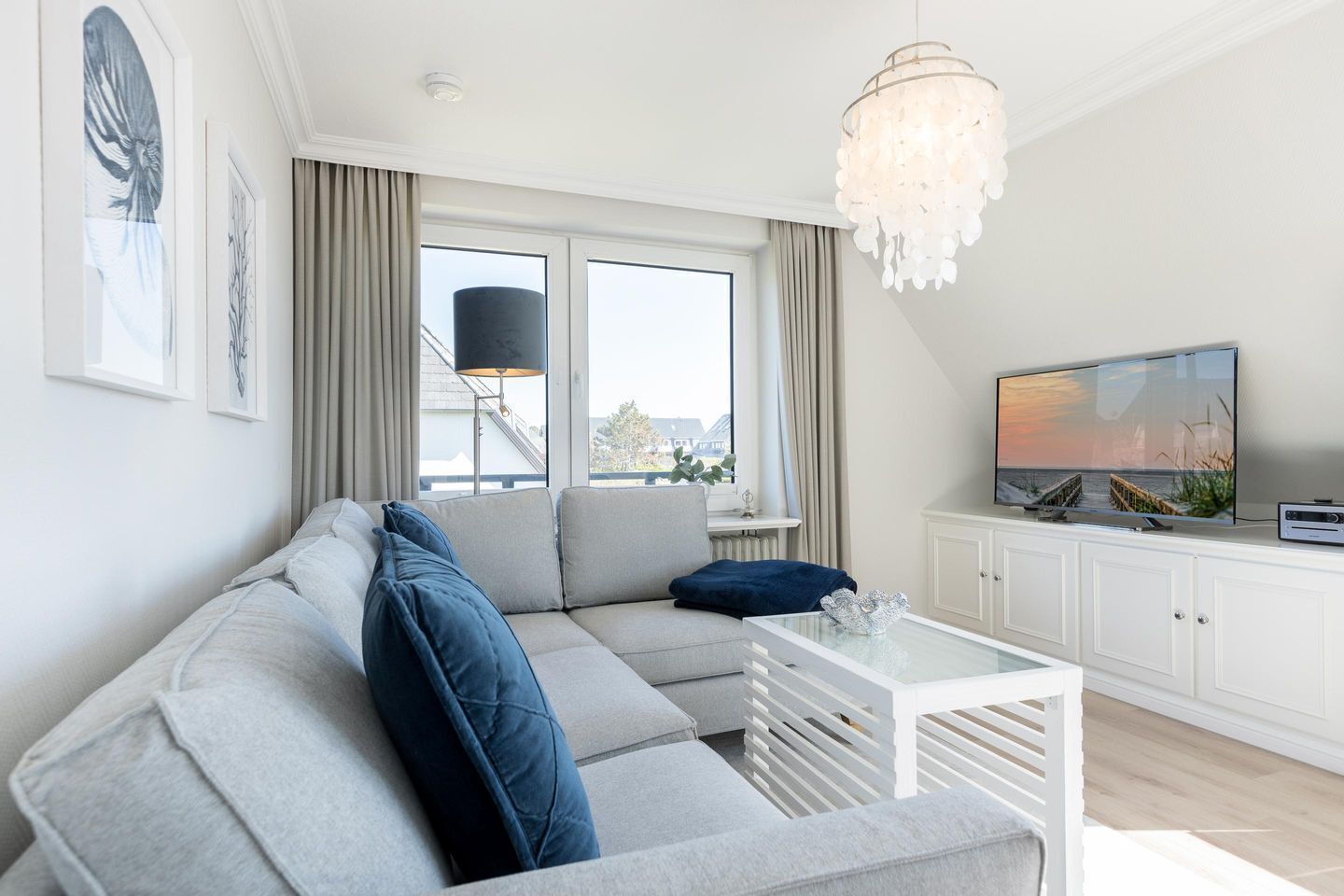 A modern living room with a grey sectional sofa, a white coffee table, a flat-screen TV, and a large window with curtains.