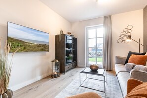 Ett modernt vardagsrum med en stor platt-TV, en bokhylla, ett soffbord med en dekorativ skål, en soffa med kuddar och ett stort fönster med utsikt över omgivningen.