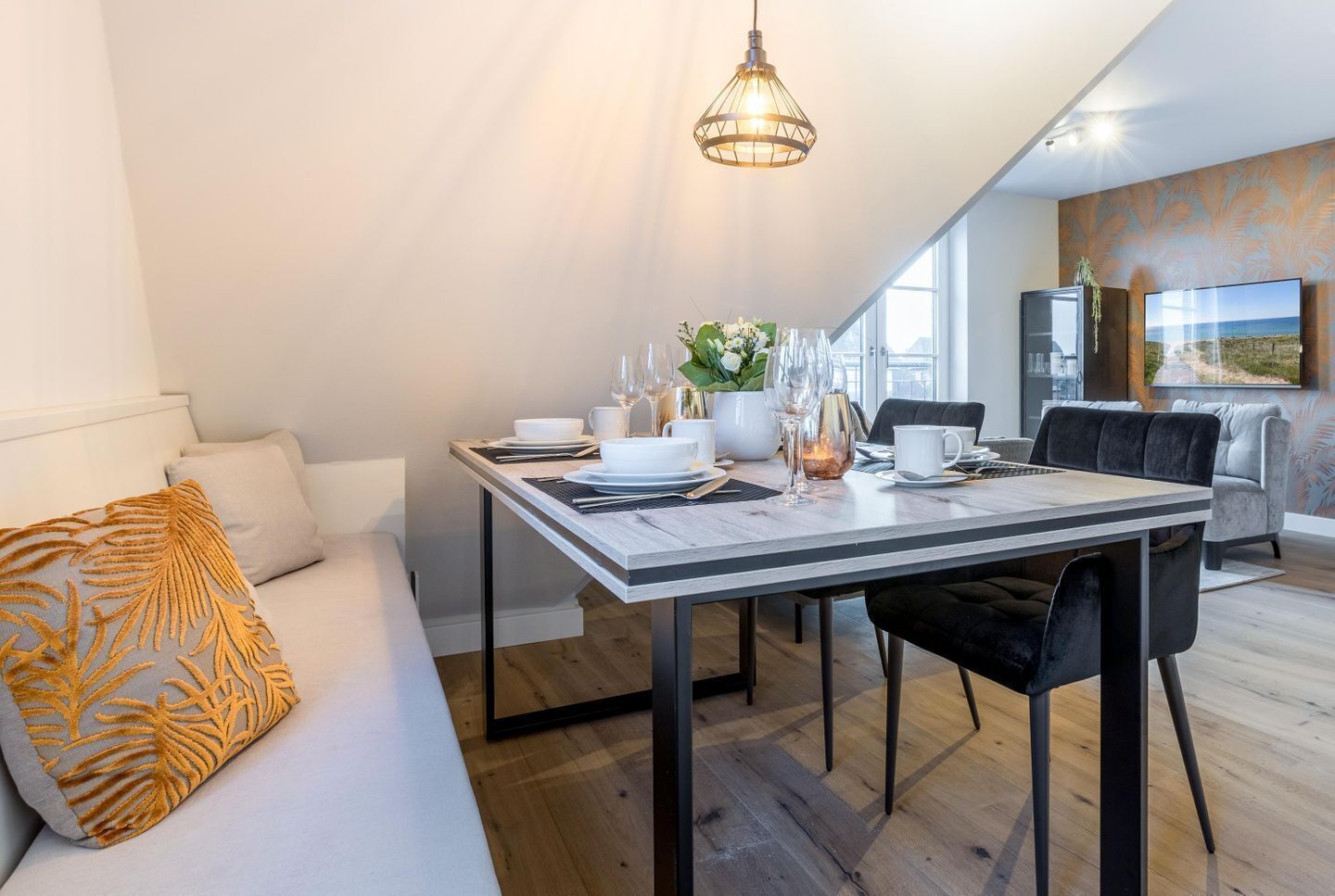 Een moderne eethoek met een gedekte tafel voor twee, een bank met een gestreept kussen, en een groot raam dat natuurlijk licht binnenlaat.