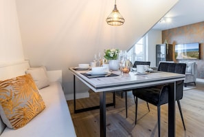 Un espace de repas moderne avec une table dressée pour deux personnes, un canapé agrémenté d’un coussin à motifs et une grande fenêtre laissant entrer la lumière naturelle.