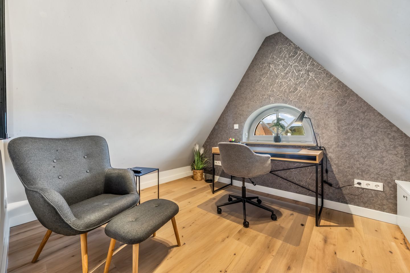 Un dormitorio tipo loft moderno con techo inclinado, un escritorio con computadora, un sillón gris y una mesita de noche pequeña.