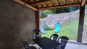 An outdoor covered patio with a table and chairs, a view of a garden, and a stone wall in the background. - Guest house "Between river and nature (Juvinas)