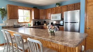 A kitchen with wooden cabinets, a stainless steel refrigerator, a microwave, and a granite countertop. There are three bar stools and a potted plant on the counter.