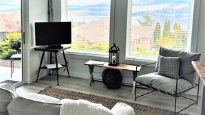 A living room with a TV on a stand, a sofa, a table with a lantern, and large windows with a view of trees and buildings.