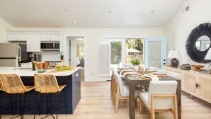 A modern kitchen with a central island, bar stools, and a dining area with a wooden table and chairs. - Sawyer by AvantStay | Stunning Isle Of Palms Home w/ Pool! (Isle Of Palms)