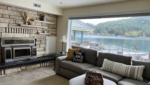 Una sala de estar con chimenea de piedra, un sofá con una almohada que dice 'NO LAKE TIME', y vista a un lago.
