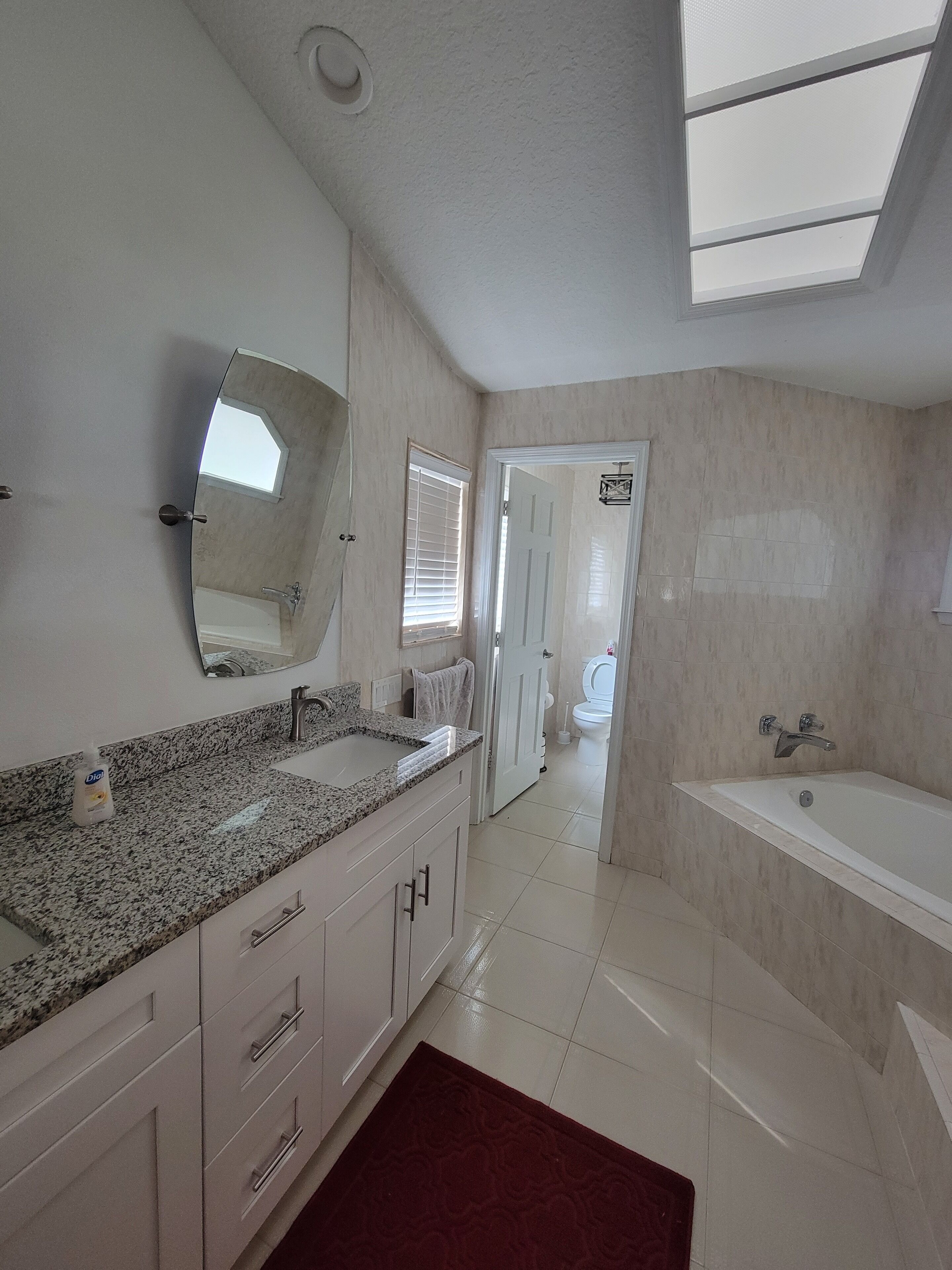 Een moderne badkamer met een granieten aanrechtblad, een grote spiegel, een bad en een toilet dat door een geopende deur zichtbaar is.