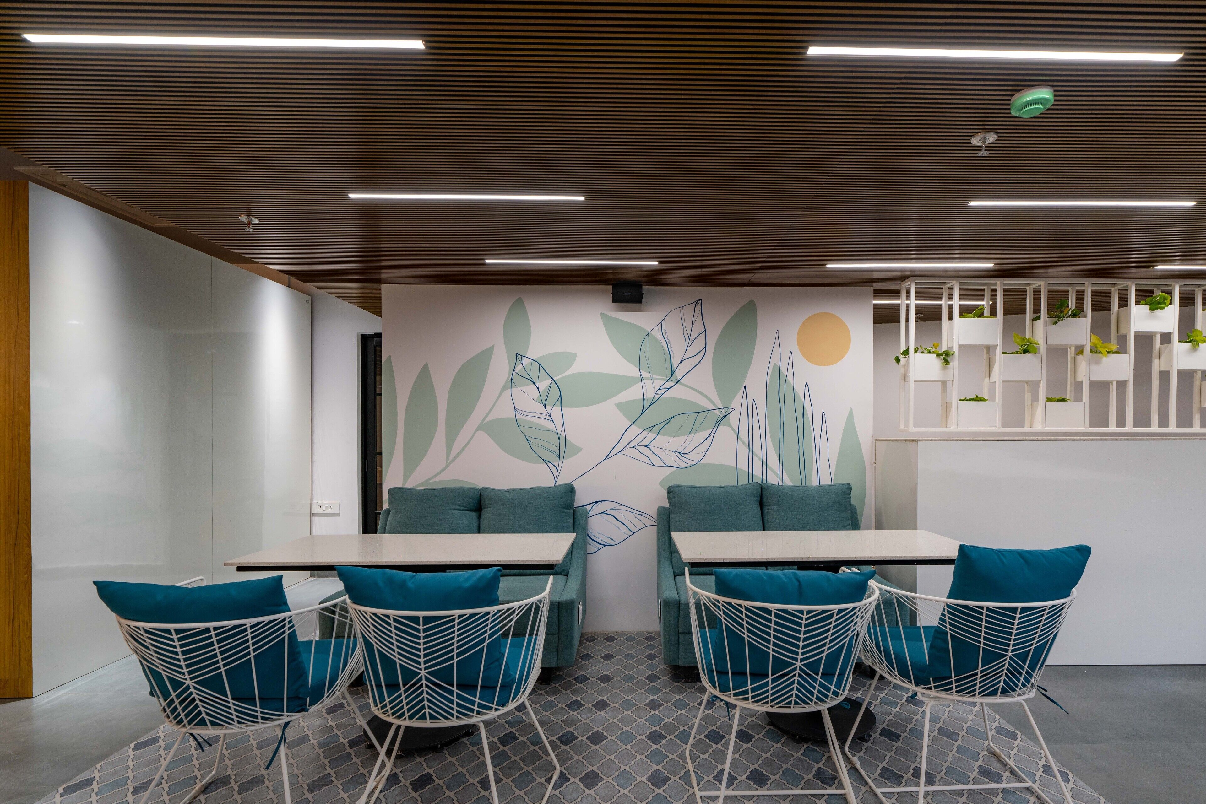 Une salle de réunion moderne avec un plafond en bois, des murs blancs et une fresque représentant des feuilles. On y trouve quatre chaises blanches avec des coussins bleus et une table.