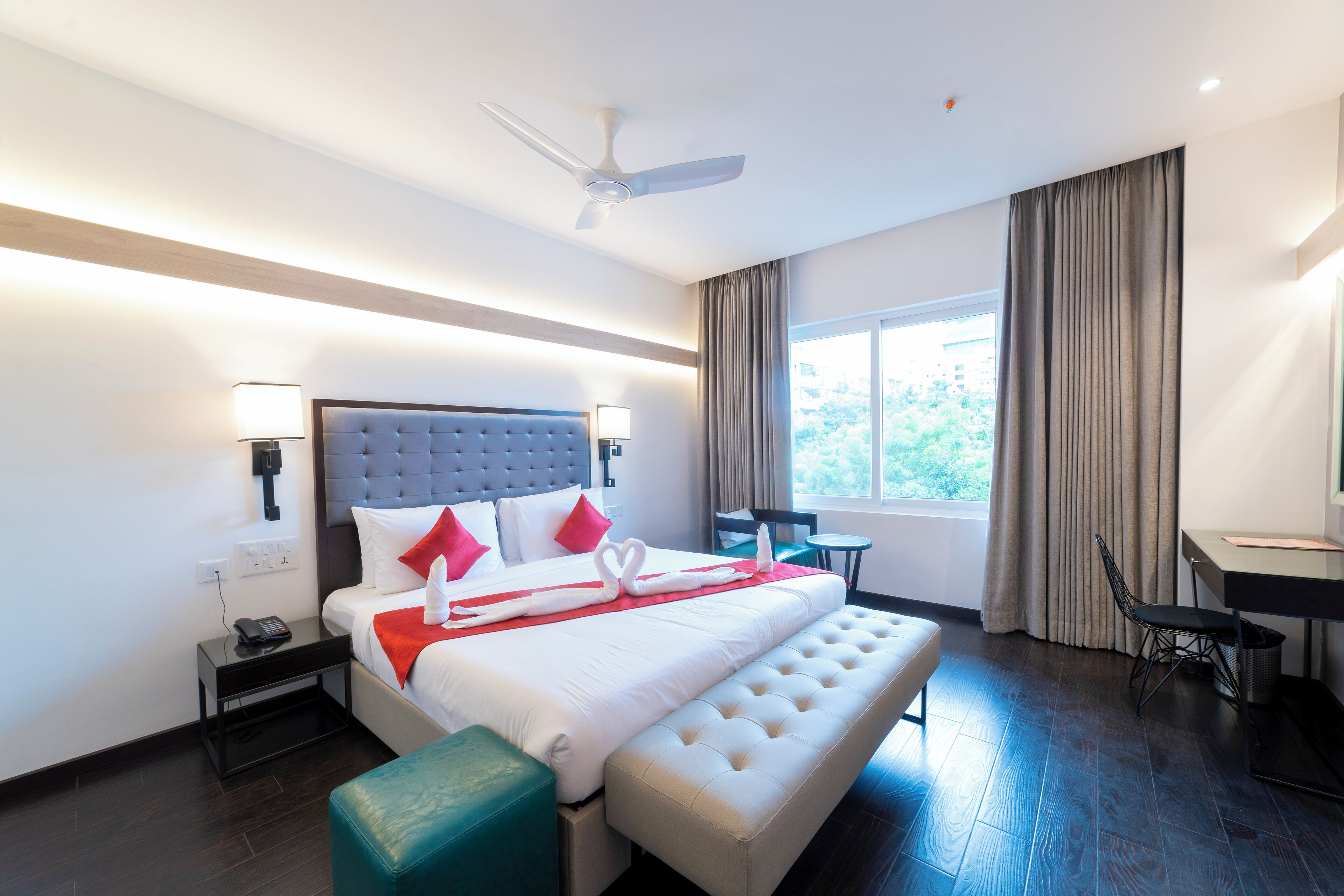 A modern hotel room with a large bed, a desk, a chair, a bench, a ceiling fan, and a window with curtains.