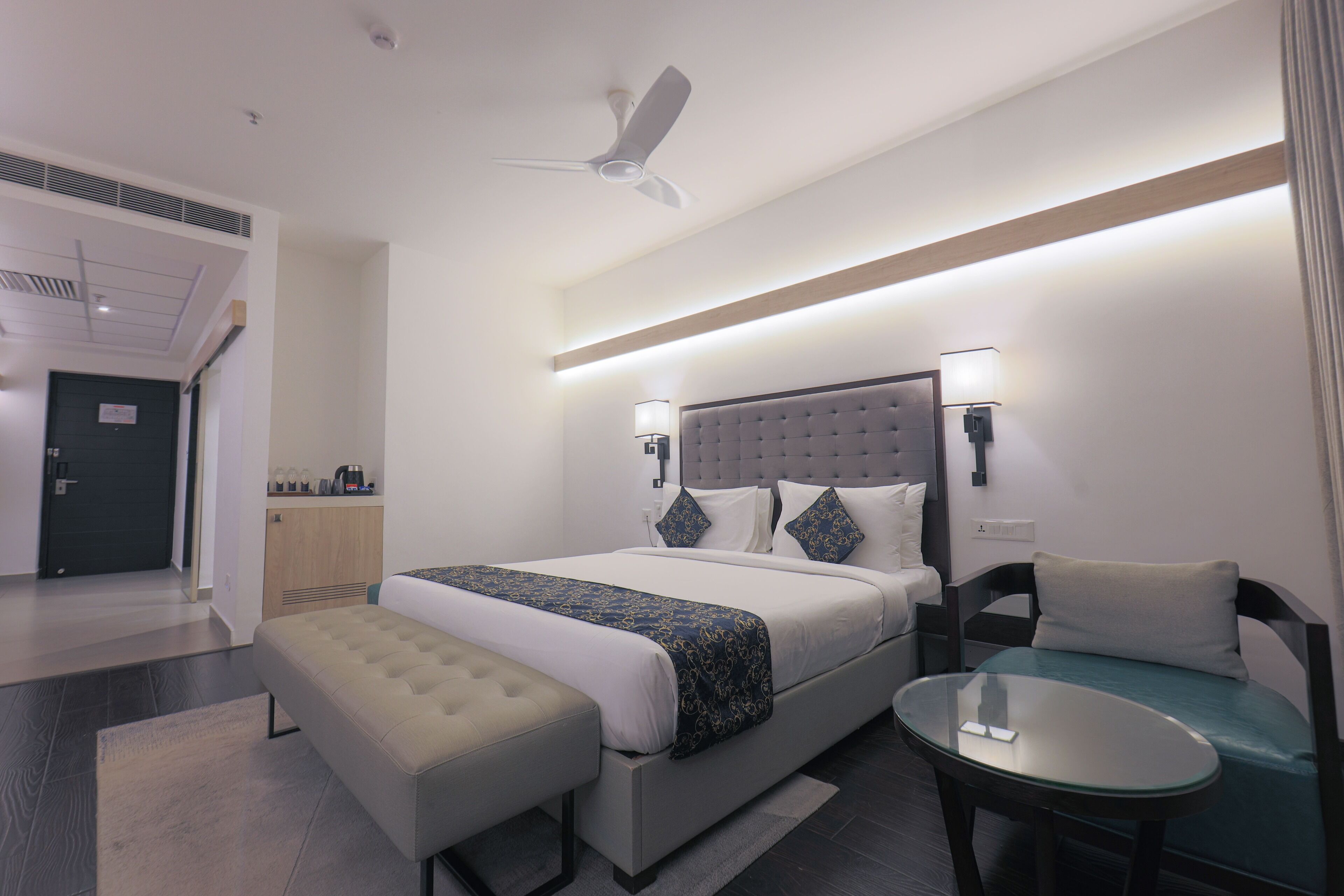 A modern hotel room with a large bed, a small table, a bench, and a ceiling fan.