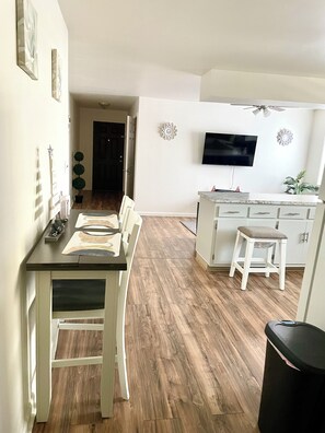 A modern kitchen with a dining area, wooden flooring, white cabinetry, a flat-screen TV mounted on the wall, and a ceiling fan.