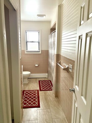 A narrow bathroom with a toilet, a shower, and a window. There are two red and white patterned rugs on the floor. - Spacious comfort, 1st FL, parking, 2 TVs, laundry,Beautiful views of NYC skyline (Clifton)