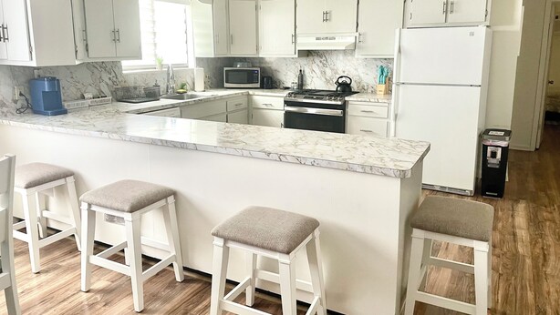 A modern kitchen with white cabinets, a marble countertop, a stove, a microwave, and a refrigerator. There are three bar stools with cushioned seats. - Spacious comfort, 1st FL, parking, 2 TVs, laundry,Beautiful views of NYC skyline (Clifton)