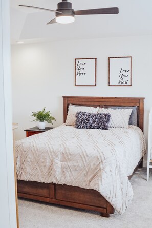 A bedroom with a wooden bed, a ceiling fan, a bedside table with a plant, and two framed inspirational quotes on the wall.