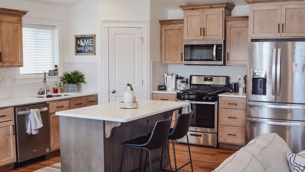 A modern kitchen with wooden cabinets, a stainless steel refrigerator, a microwave, and a stove. There is a small island with two bar stools.