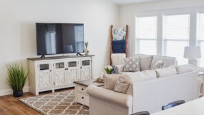 A living room with a large flat-screen TV, a white cabinet with glass doors, a beige sofa, and a patterned rug.