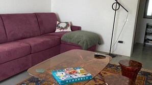 Ein Wohnzimmer mit einem violetten Sofa, einem Glastisch mit einem Scrabble-Spiel, einer Stehlampe und einem gemusterten Teppich.