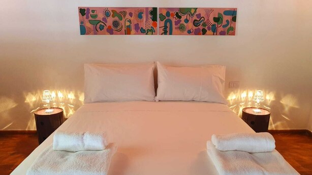 Ein ordentlich bezogenes Bett mit weißen Leinentüchern und zwei Kissen, begleitet von zwei Nachttischen mit Lampen, in einem Zimmer mit schlichter Wand und einem farbenfrohen, abstrakten Gemälde über dem Bett.