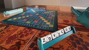 Ein Scrabble-Spiel auf einem gemusterten Teppich, im Hintergrund ein Fernseher, der ein stürmisches Meer zeigt.