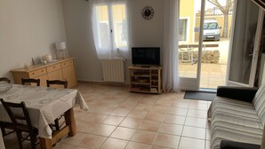 A living room with a sofa, a TV on a wooden stand, a dining table with chairs, a wooden cabinet, and a window with curtains. - Gîte Caramolle, St Denis, Gard, Cévennes, 2 bedrooms, swimming pool, nature (Saint-Denis)