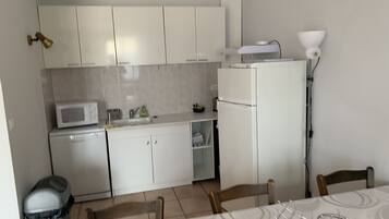 Une cuisine compacte avec des meubles blancs, un four à micro-ondes, un réfrigérateur et un évier. On y trouve une table à manger avec des chaises et une poubelle.
