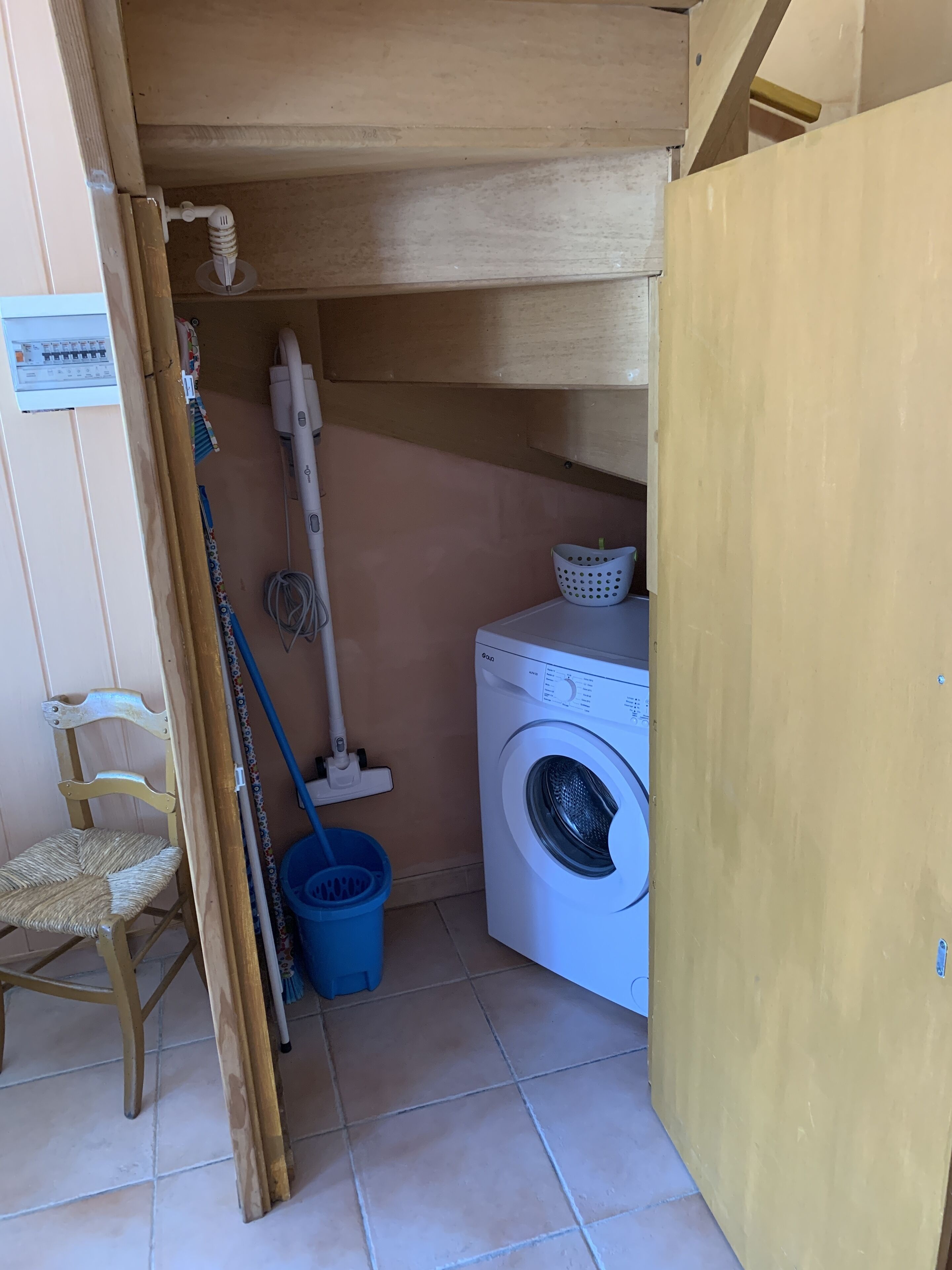 A laundry room with a washing machine, a mop, a broom, a blue bucket, and a wooden chair.
