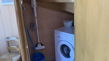 A laundry room with a washing machine, a mop, a broom, a blue bucket, and a wooden chair.