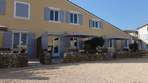 A building with a yellow facade, multiple windows with shutters, outdoor seating with umbrellas, and a gravel ground. - Gîte Caramolle, St Denis, Gard, Cévennes, 2 bedrooms, swimming pool, nature (Saint-Denis)