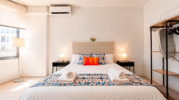 A hotel room with a bed, two bedside tables, a window, an air conditioning unit, and a shoe rack.