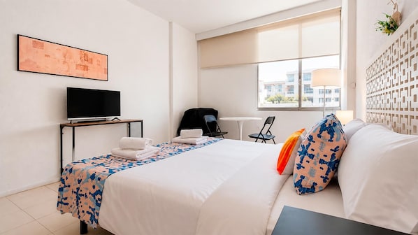 Quarto de hotel com cama, escrivaninha, cadeira, televisão e janela com vista para edifícios.