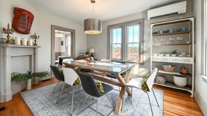 Un coin repas avec une table en bois, des chaises et un lustre. On y trouve une cheminée agrémentée d’objets décoratifs et d’une plante.