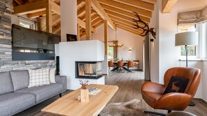 A modern living room with a stone fireplace, wooden beams, a leather armchair, and a dining area in the background. - Luxurious chalet with sauna, near the ski slope (Turracher Höhe)