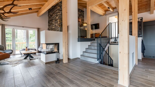 A spacious living area with a stone fireplace, a staircase, and a large window overlooking a deck. - Luxurious chalet with sauna, near the ski slope (Turracher Höhe)