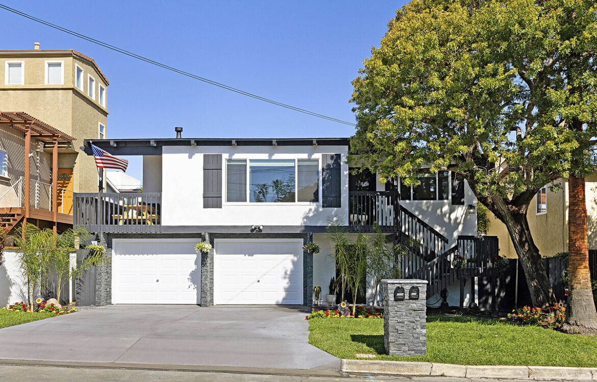 Une maison moderne à deux étages, avec une porte de garage blanche, un balcon doté d’une balustrade et un grand arbre dans le jardin avant.