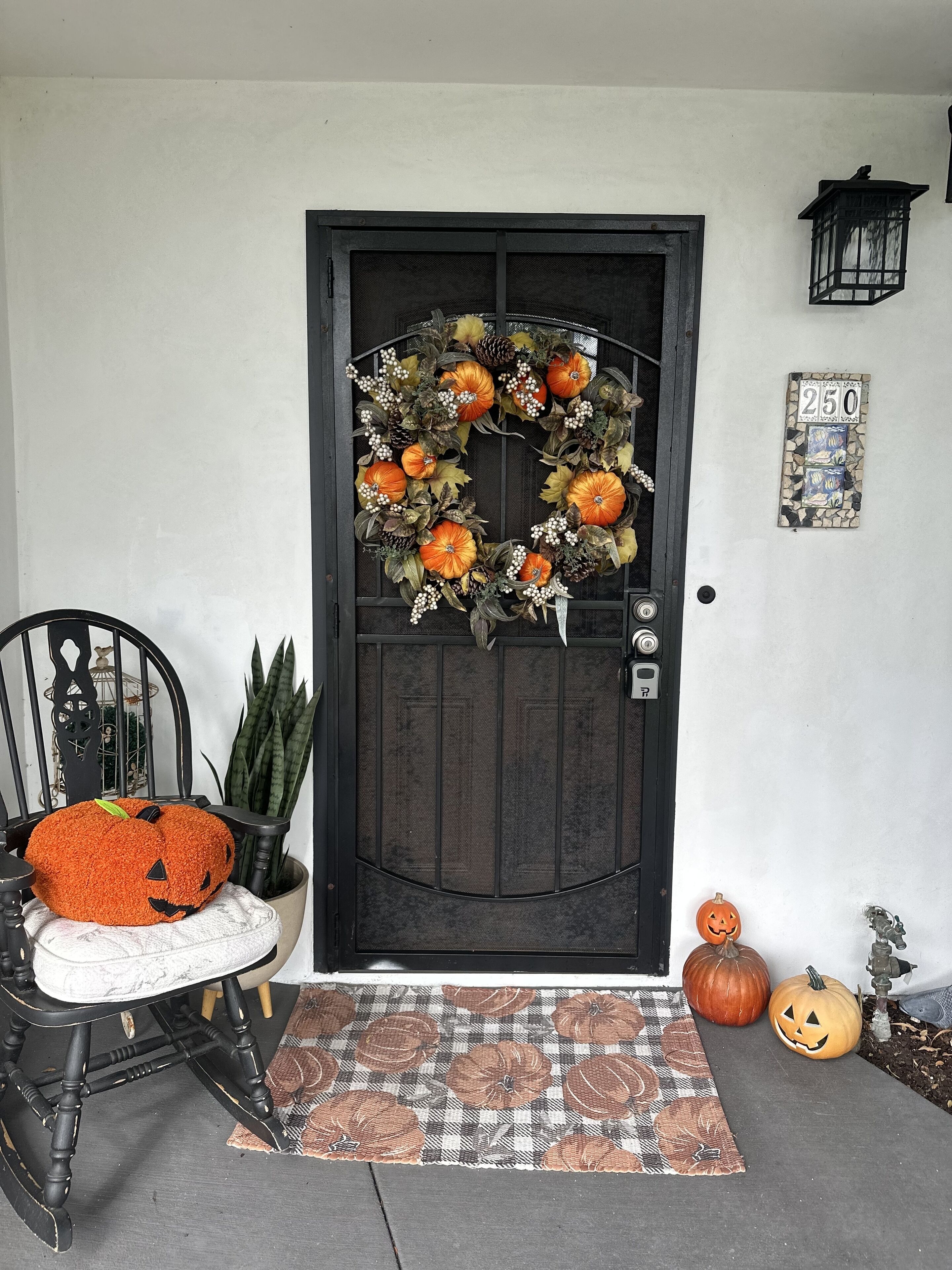 A dark wooden door with a wreath, a chair with a pumpkin cushion, and two pumpkins on the ground.