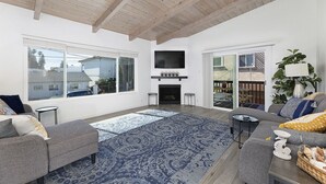 A spacious living room with a fireplace, large windows, and a ceiling with exposed beams. - Cheerful and Remodeled 3-bedroom beach cottage, STEPS to beach. Sleeps 9!  (Carlsbad)