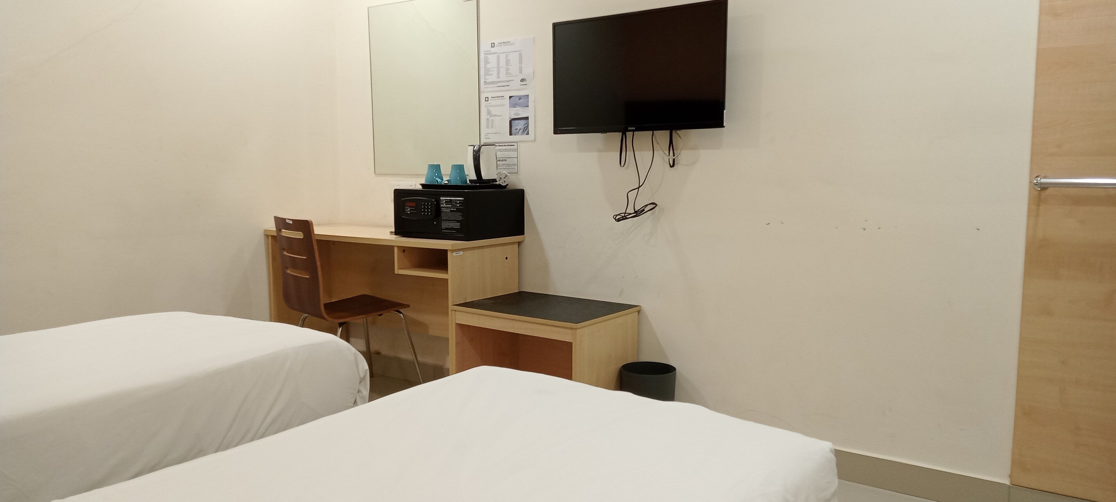 Une chambre d’hôtel avec deux lits, un bureau avec un ordinateur, un téléviseur fixé au mur et une poubelle.