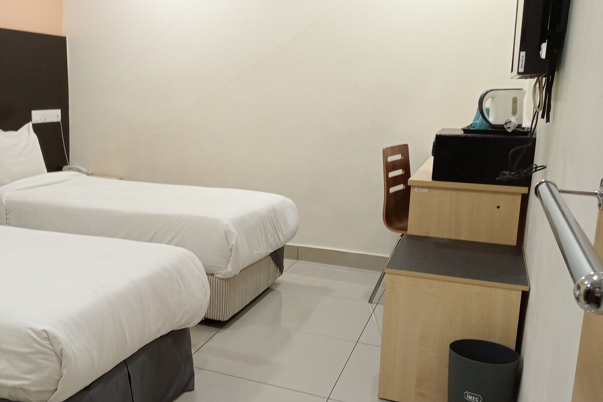 A hotel room with two beds, a desk, a chair, a trash bin, and a wall-mounted TV.
