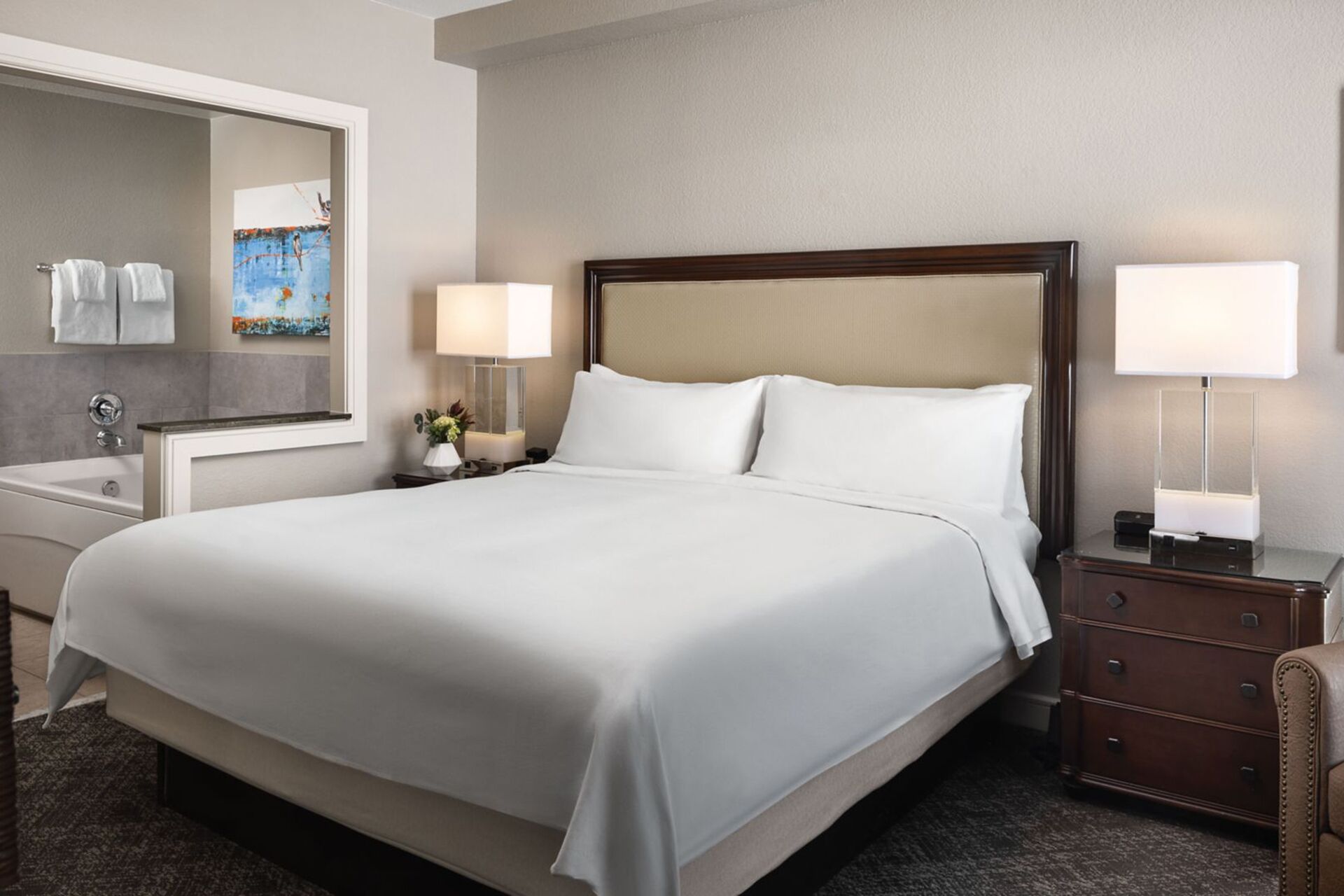 A hotel room with a large bed, a bedside table with a lamp, a dresser, a mirror, and a painting on the wall.
