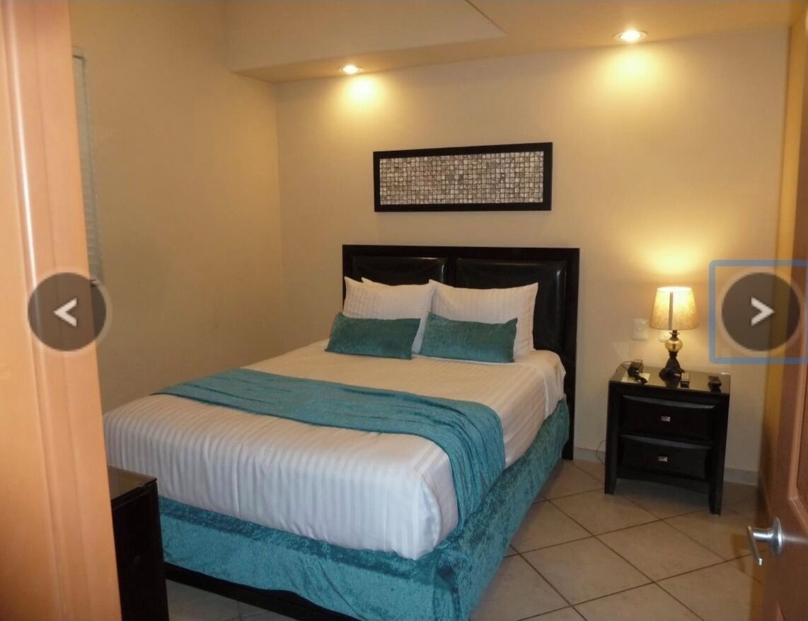 A neatly made bed with a teal bedspread and pillows, a bedside table with a lamp, and a framed artwork on the wall.