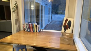 Ein Holztisch mit Büchern, einem Hahn-Gemälde und einem Fenster mit Blick auf ein Haus und Berge.
