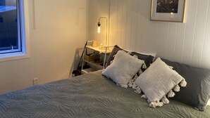 Ein Schlafzimmer mit einem Bett, Kissen, einem Nachttisch mit Lampe, einem gerahmten Bild an der Wand und einem Fenster.