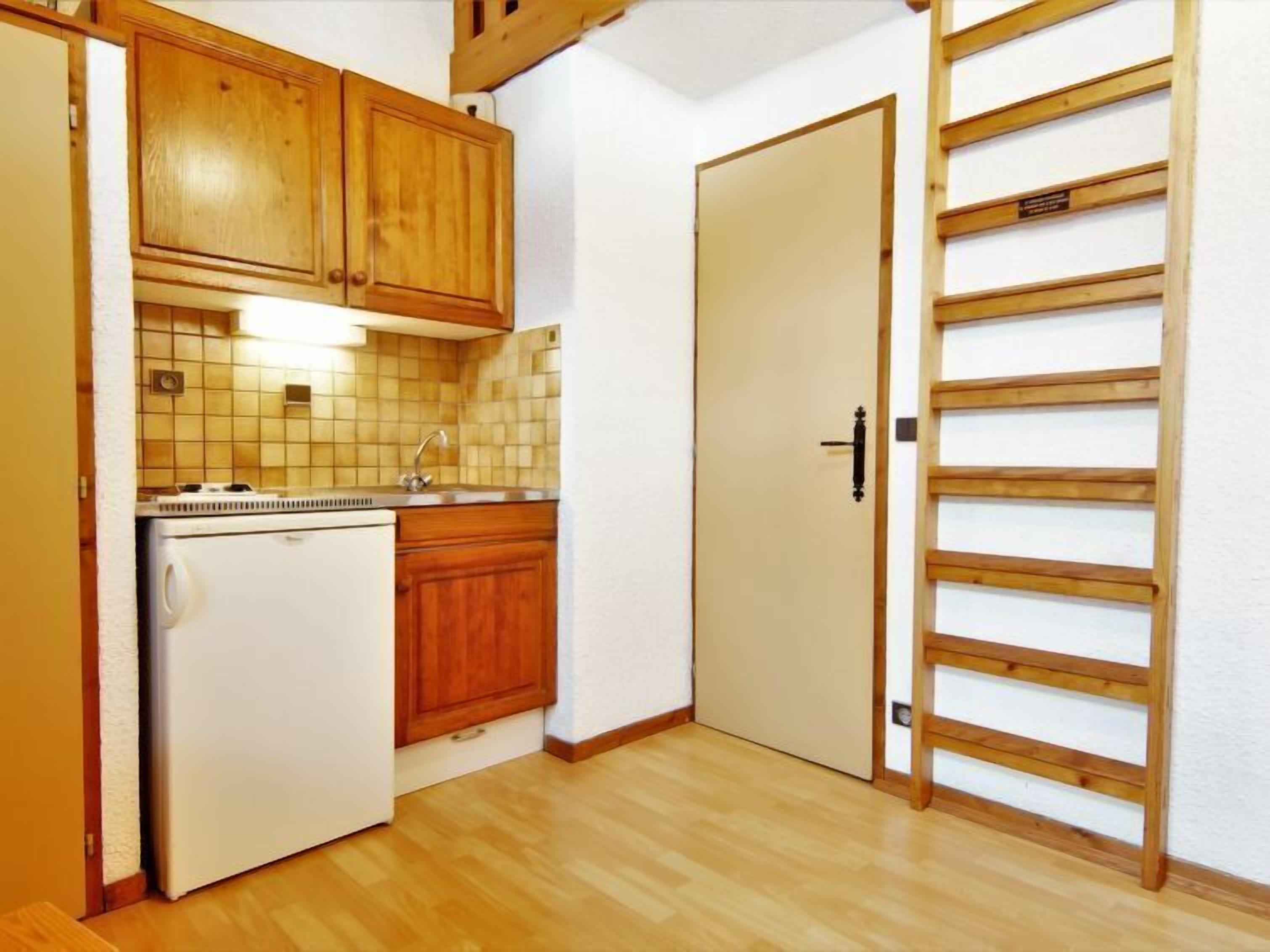 Eine kompakte Küche mit Holdekor-Schränken, einer gefliesten Spritzschutzleiste, einem eingebauten Kühlschrank und einem Holzleiterregal.