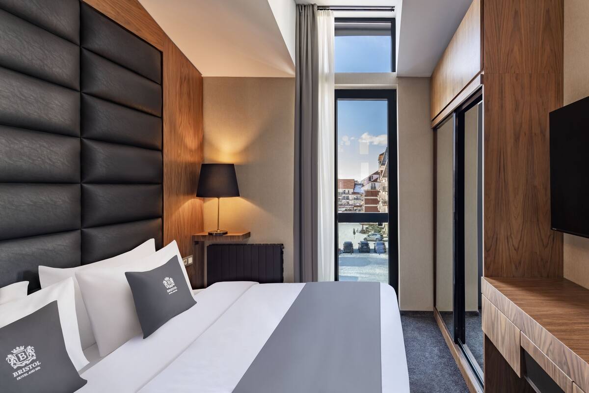 A modern hotel room with a large bed, a wooden headboard, a bedside table with a lamp, and a view of the city through a large window.