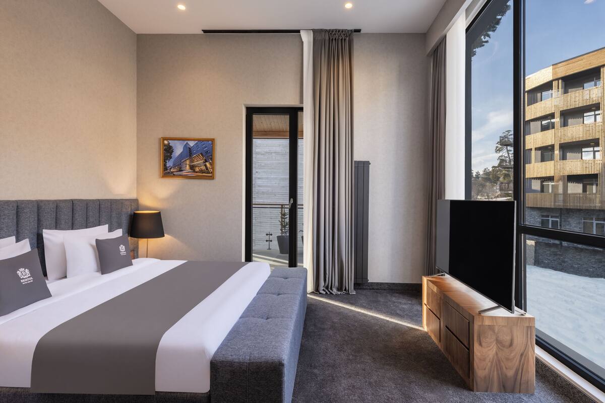 A modern hotel room with a large bed, a TV, a balcony, and a view of the city.