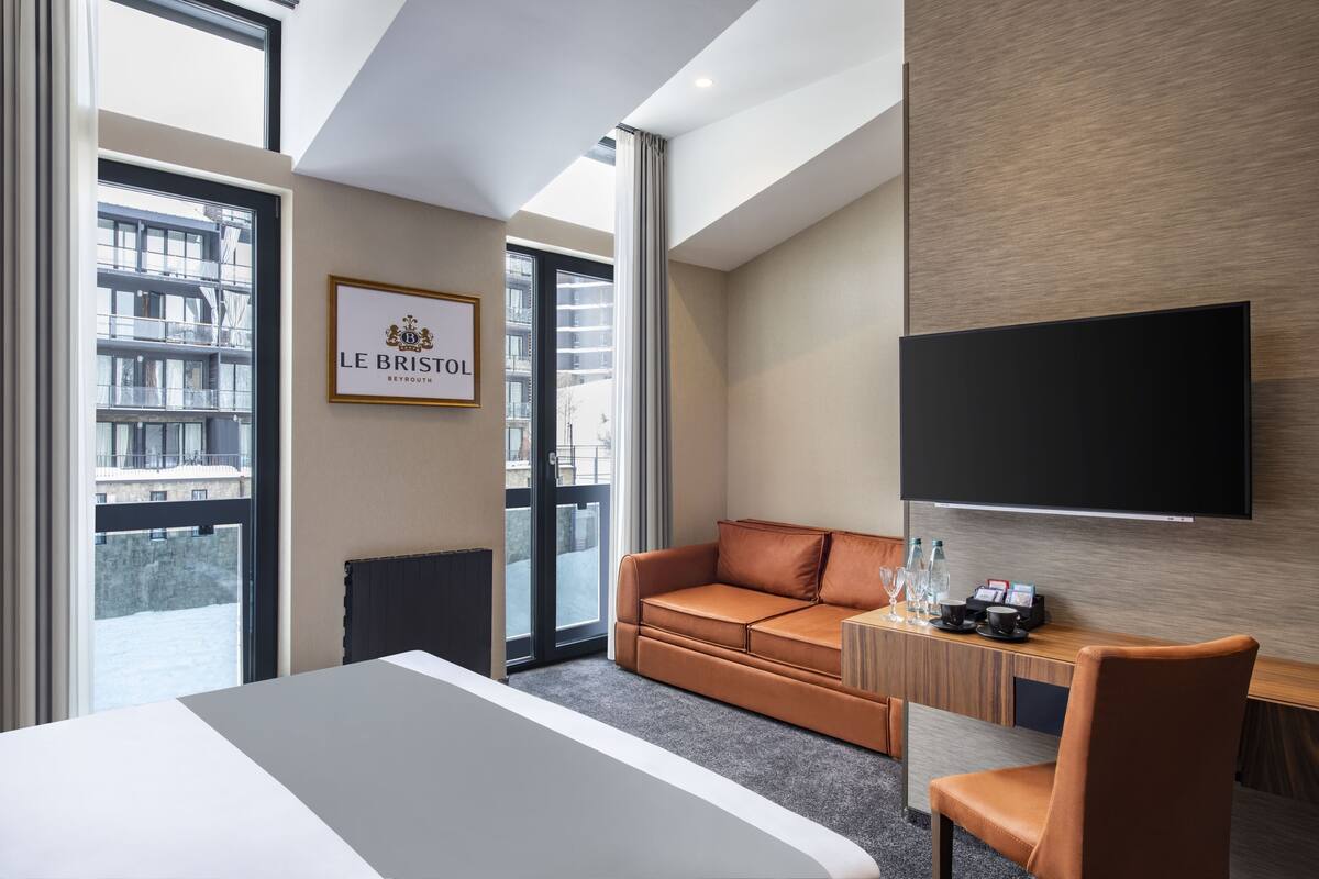 A hotel room with a large bed, a sofa, a desk with a flat-screen TV, and a window with a view of buildings.