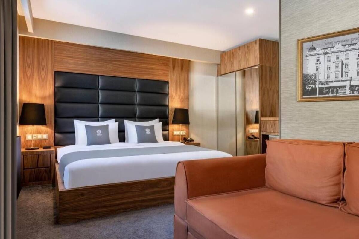 A modern hotel room with a large bed, wooden headboard, two bedside tables with lamps, a wardrobe, and a framed picture on the wall.