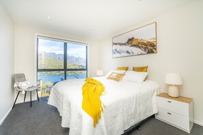A bedroom with a large window, a bed with a white and yellow bedspread, a bedside table with a lamp, and a chair.